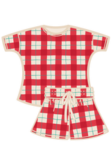 A two-piece women's pyjama set featuring a short-sleeve top and matching shorts in a red, white, and green tartan, with a cream-colored collar, pocket, and drawstring. Made from organic cotton